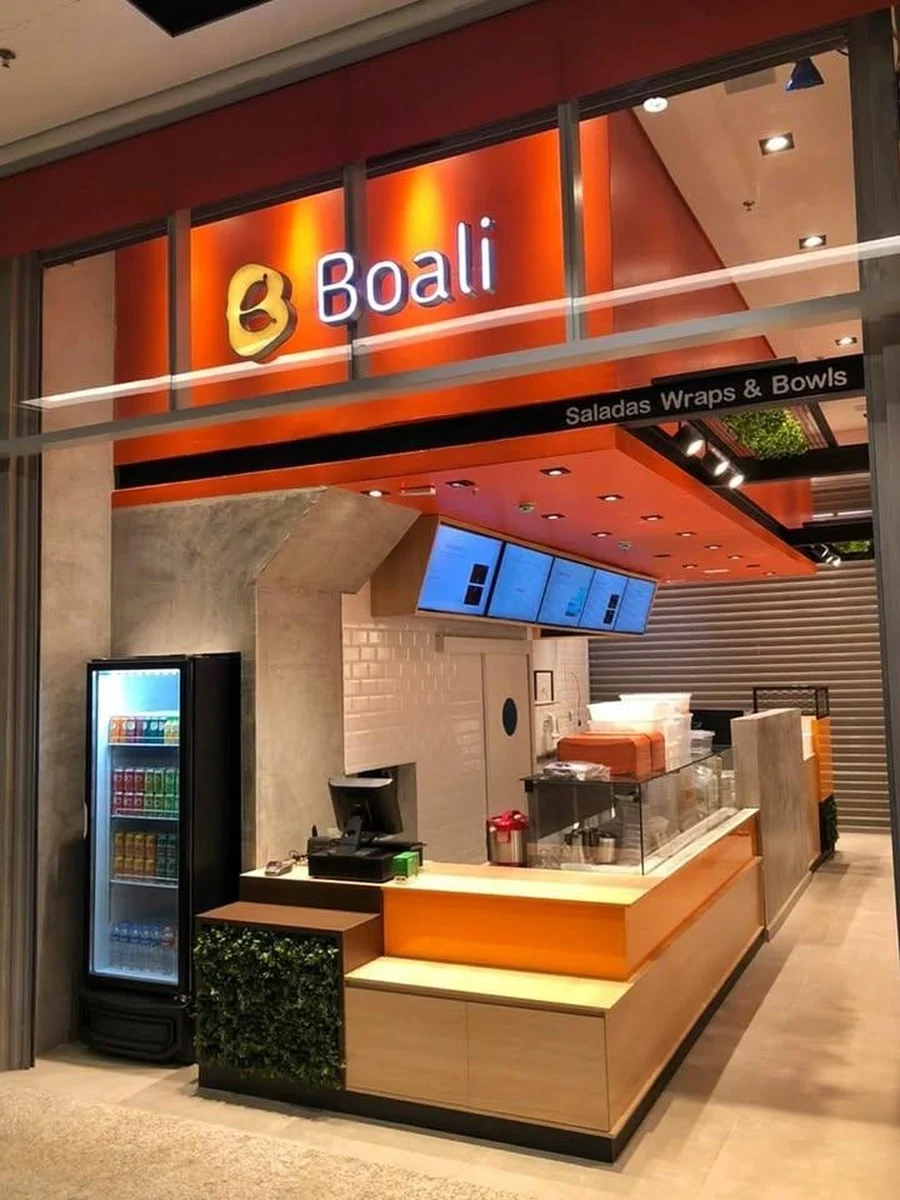 Fachada de loja em shopping, com letreiro iluminado, balcão de atendimento em madeira clara e expositor refrigerado.